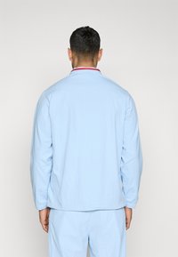 Chaqueta azul claro con manga larga, puños elásticos y un collar de acento rojo. Hecha de tela suave, diseñada para un ajuste relajado.