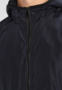 Svart zip-up-jacka med hög krage som är gjord av ett slätt, lätt material. Har en elegant design, minimal sömnad och inga synliga mönster.