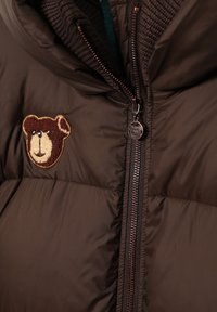 Barna pehelydzseki puha mackóemblémával, barna cipzárral és bordázott gallérral. Simaságot és párnázott textúrát kínál, amely meleget és szigetelést biztosít.