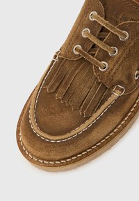 Καφέ μοκασίνι από σουέντ με φουντάκια, λευκή ραφή και στρογγυλά κορδόνια περασμένα από μεταλλικά μπούτια σε ανοιχτό φόντο.