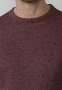 Primo piano di un uomo che indossa un maglione girocollo bordeaux realizzato in un tessuto leggermente testurizzato, con un piccolo logo ricamato sul petto.