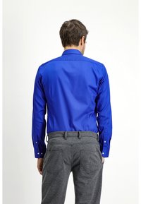 Camisa azul de manga larga con botones, con un cuello clásico y botones en los puños, combinada con pantalones grises texturizados, vista desde la parte de atrás.