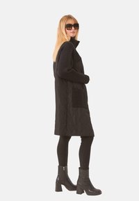 Abrigo negro acolchado con mangas de punto trenzado, bolsillos laterales, cuello alto, combinado con leggings negros y botas de tobillo. Textura suave y diseño minimalista.