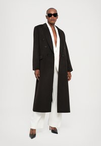 Lang, dubbelzijdig zwart wollen jas met aangeknipte revers, voorzien van grote zwarte knopen en een gladde textuur, gedragen over een witte blazer en broek.