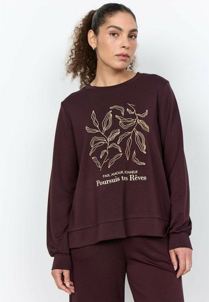 Vrouw met krullend haar in een paardenstaart, gekleed in een donkerpaarse trui met een beige bladdesign en Franse tekst "Poursuis tes Rêves".