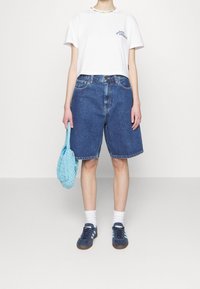 Camiseta blanca recortada con logo, pantalones cortos de mezclilla azul, bolso tejido azul, zapatillas deportivas azules con rayas azul claro, calcetines blancos hasta el tobillo.