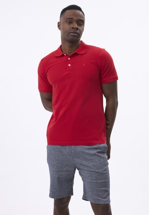 Man staat met handen achter de rug, draagt een rood poloshirt en grijze korte broek tegen een witte achtergrond.