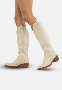 Botas de vaquero beige con un caña alta, cremallera lateral y bordado de hojas en relieve. Tacón grueso marrón y textura de cuero suave.