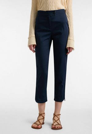Donna che indossa pantaloni navy corti con spacchi laterali, maglione beige a trecce con polsini estesi e sandali marroni con cinturini.