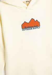 Kr�ämfärgad hoodie med känguruficka och broderad bergdesign i orange och blått, med texten "OUTDOOR SUPPLY."