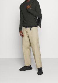 Pull en tricot vert foncé, pantalon beige à coupe décontractée et baskets noires. L'ensemble comprend un sac à bandoulière noir avec des zips et un détail ceinture.