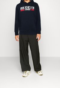 Felpa blu navy con logo "JACK & JONES" in rosso e bianco, dotata di tasca a canguro. Abbinata a pantaloni neri larghi e scarpe da ginnastica bianche.
