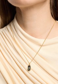 Collar de oro con un colgante ovalado oscuro, reposando sobre una tela plisada de un beige claro. Textura suave y diseño simple y elegante.