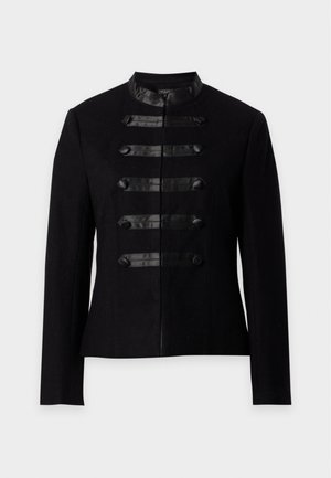Femme debout, mains dans les poches, portant une veste noire de style militaire, un haut noir, un jean large bleu foncé et des chaussures noires.