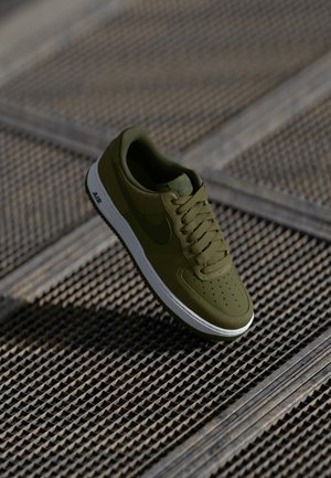 Sneaker Nike Air vert olive avec semelle blanche, montrée en train de flotter au-dessus d’une surface de grille métallique texturée.