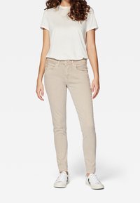 Beige skinny jeans with five pockets, button closure, and stitched detailing, paired with a white short-sleeve t-shirt and white sneakers.