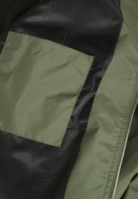 Olive green fabric with a black satin panel, featuring a rectangular patch pocket, smooth texture, and contrasting zipper detail.