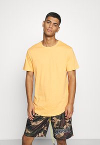 Oranges Baumwoll-T-Shirt mit Rundhalsausschnitt und kurzen Ärmeln, kombiniert mit kurz geschnittenen Shorts im Camouflage-Muster, die ein Logo in der Nähe des Saums aufweisen.