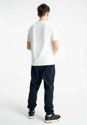 Młody mężczyzna stoi tyłem, ubrany w białą koszulkę, ciemnogranatowe dresy i czarne trampki z białymi podeszwami.