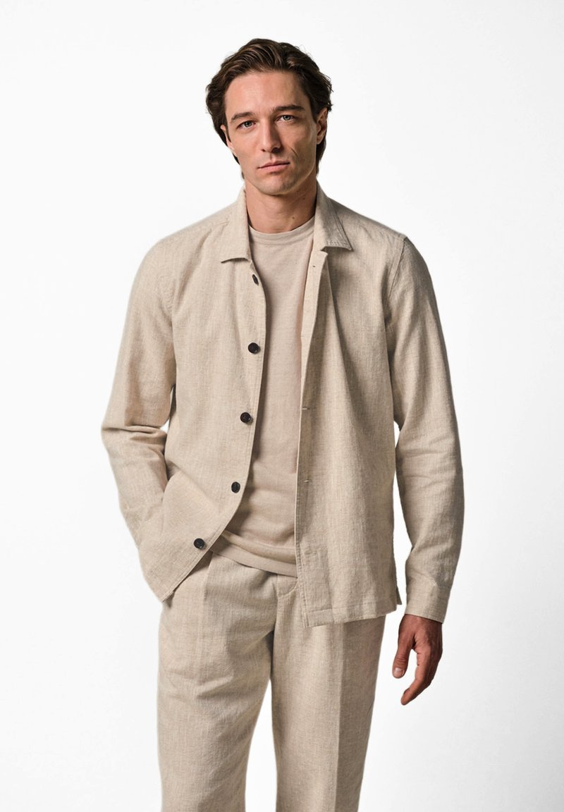 Man wearing beige button-up shirt over matching T-shirt and beige pants, standing with one hand in pocket against white background.