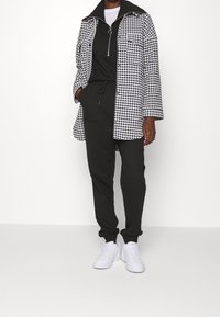 Manteau long en pied-de-poule noir et blanc porté sur un haut zippé noir et un jogging, avec des baskets blanches. Tissu lisse, coupe décontractée, deux poches.