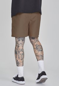 Shorts marron avec une texture lisse, portés avec des chaussettes blanches et des baskets noires. Des tatouages détaillés sur les jambes présentent divers motifs et portraits.