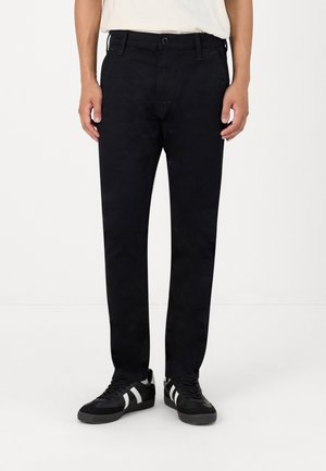 Mann trägt schwarze Slim-Fit-Hose, schwarze Sneaker mit weißen Streifen und ein weißes, in die Hose gestecktes Hemd und steht vor einem weißen Hintergrund.