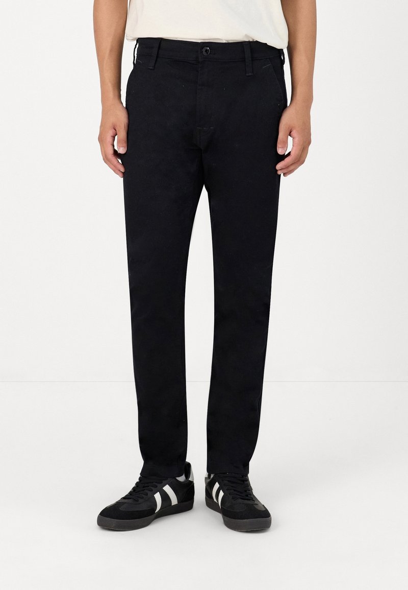 Homme portant un pantalon noir slim, des baskets noires avec des bandes blanches et une chemise blanche rentrée, debout devant un fond blanc.