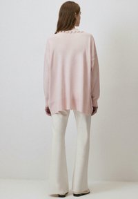 Pulover oversized de culoare roz deschis, cu tiv ondulat și embellisment pe spate. Asortat cu pantaloni evazați de culoare crem, creând un aspect stratificat.