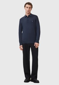 Marineblaues Langarm-Polohemd mit kragen, drei Knöpfen und einem dezenten Emblem. Kombiniert mit schwarzen Hosen und Schuhen.