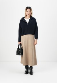 Femme se tenant contre un fond blanc, portant une veste zippée bleu marine, une longue jupe beige, des bottes noires, tenant un sac à main noir dans sa main gauche.