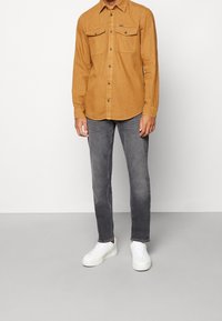 Brązowa koszula zapinana na guziki z dwoma kieszeniami na piersi, w połączeniu z ciemnoszarymi jeansami i białymi trampkami. Czysty design, dopasowany fason i gładka tekstura.