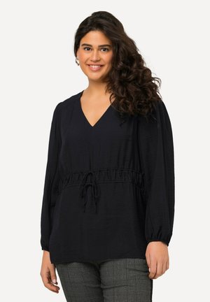 Blusa nera in tessuto strutturato con scollo a V, vita elastica e maniche lunghe a sbuffo. Indossata sopra pantaloni fantasia.