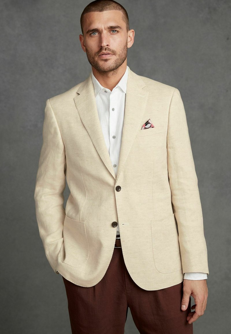Blazer en lin beige avec deux poches avant, boutons marron, et un carré de poche présentant des motifs géométriques, associé à une chemise blanche et un pantalon marron.