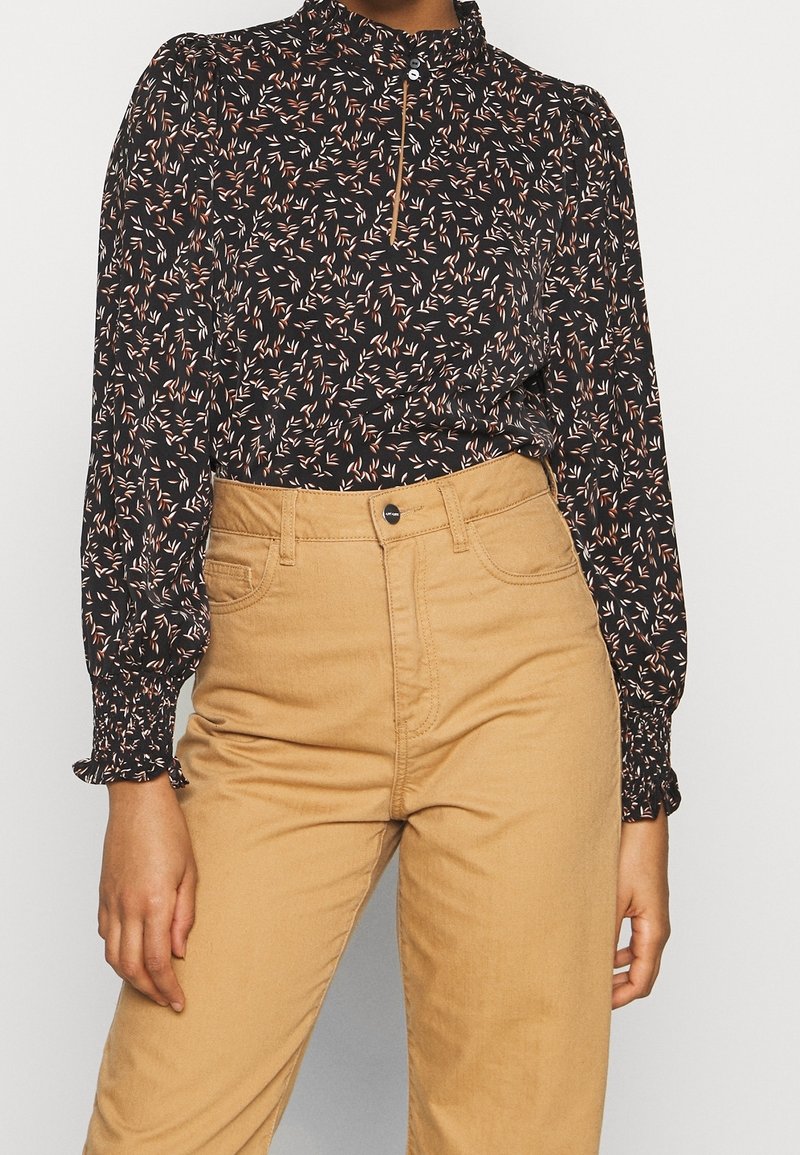 Femme portant un chemisier noir à manches longues avec un petit motif de feuilles et un pantalon beige taille haute, mains détendues le long du corps.