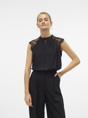Chemisier noir avec des épaules accentuées de dentelle, décolleté en trou de serrure, tombé en tissu doux, associé à un pantalon noir taille haute, le modèle pose de manière neutre.