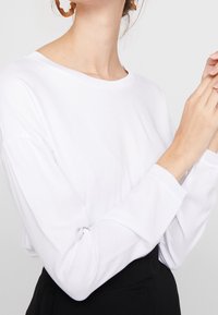 Vit långärmad topp med rund halsringning, tillverkad av mjukt tyg. Modellen bär en svart kjol, med händerna ihop, och stora bruna örhängen.