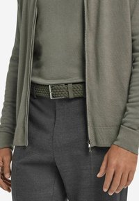 Olivgrön zip-up jacka över en matchande topp, i kombination med grå byxor och ett flätat grönt bälte med en silverfärgad spänne.