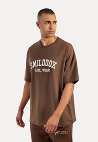 Camiseta oversized marrón hecha de un tejido suave que presenta "SMILODOX ATHL. WEAR" impreso en blanco. Diseño de cuello redondo y corte relajado.