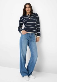 Top a righe blu navy lavorato a maglia con tre bottoni, abbinato a jeans a gamba larga azzurri e sneakers bianche, su uno sfondo neutro.