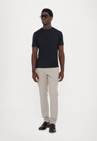 Dunkelblauses Baumwoll-T-Shirt, Slim Fit, kombiniert mit hellbeigen, maßgeschneiderten Hosen. Schwarze Slipper runden das Outfit ab. Model steht vor einem weißen Hintergrund.