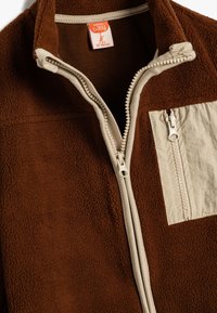 Chaqueta de forro marrón con acentos en beige. Presenta un alto cuello, cierre de cremallera completo y un bolsillo en el pecho con cremallera. Textura suave.
