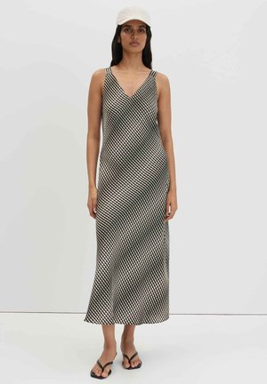 Femme portant une robe sans manches à motif diagonal noir et blanc, une casquette blanche et des sandales slides noires, debout devant un fond clair uni.