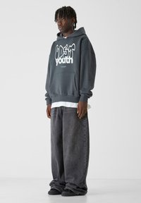 Felpa grigia antracite con vestibilità oversize, caratterizzata da testo grafico bianco. Abbinata a jeans in denim scuri a vestibilità larga e scarpe da ginnastica nere.