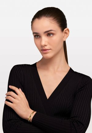 Brățară din aur pe un încheietură, purtată cu un top lung cu mâneci bufante de culoare neagră, având un design adânc în V, evidențiind contrastul de textură și material.