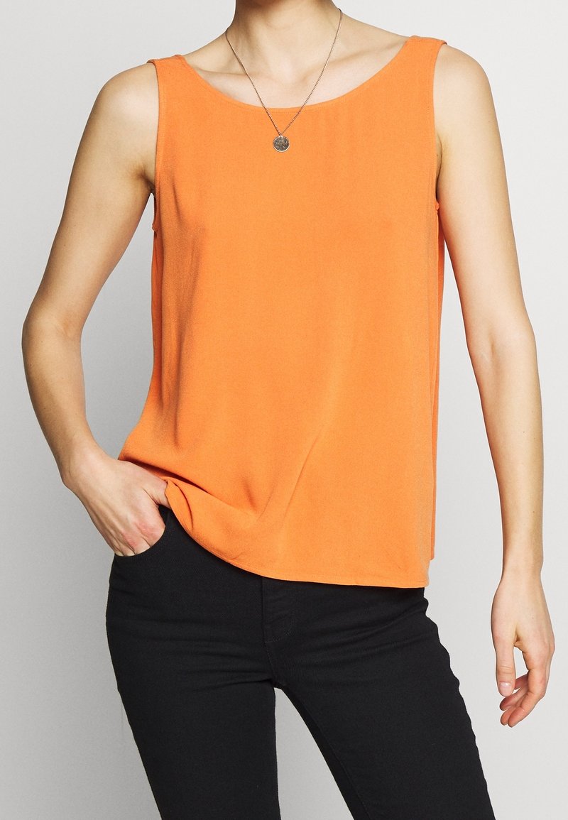 Haut sans manches orange vif porté avec un pantalon noir, accessoirisé d’un petit pendentif rond.
