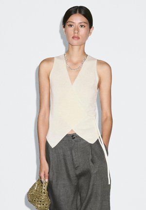 Beige Wickeloberteil mit tiefem V-Ausschnitt, strukturiertem Strickstoff und bindbarem Verschluss an der Seite, kombiniert mit grauen, locker sitzenden Hosen.