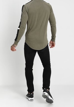 Homme portant une chemise manches longues vert olive avec des rayures noires et blanches sur les manches, un pantalon noir et des baskets noires, marchant de dos.