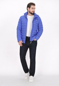 Blå puffjacka med hög krage, dragkedja och quiltad textur, kombinerad med mörka jeans och vita sneakers.