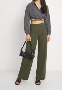 Šedý zkrácený top s nabíranými rukávy, spárovaný s vysokými olivově zelenými širokými kalhotami. Drží malou černou kabelku a má na sobě černé sandály.